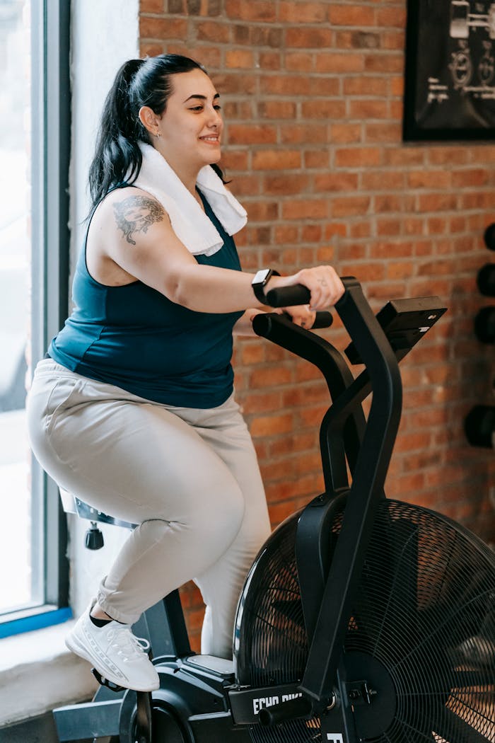 Side view of cheerful plus size ethnic female in activewear with towel exercising on fitness equipment during workout in gym